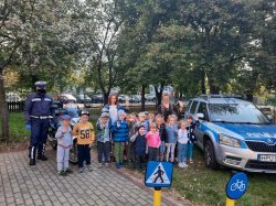 Z prawej strony stoi radiowóz policyjny, obok niego grupa dzieci w wieku przedszkolnym wraz z opiekunkami i policjantem