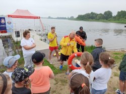 Na obrazku widzimy jezioro oraz ratowników WOPR prezentujących dzieciom koło ratunkowe