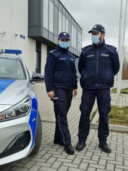 policjantka i policjant w umundurowaniu i w maseczkach  stoją przy radiowozie. W tle widać budynek komendy.