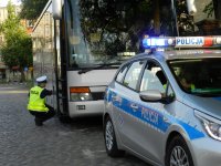 Na zdjęciu widoczny jest radiowóz policyjny za którym zaparkowany jest autobus przy którym czynności kontrolne wykonuje umundurowany policjant.