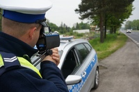 Na obrazku widzimy umundurowanego policjanta dokonującego pomiaru prędkości jadących pojazdów. Obok policjanta widzimy zaparkowany radiowóz policyjny.