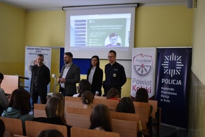 Na obrazku widoczni są: umundurowany policjant oraz Powiatowy Rzecznik Konsumentów w trakcie prowadzenia prelekcji z okazji obchodów Światowego Dnia Konsumenta.