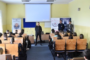 Na obrazku widoczni są: umundurowany policjant oraz Powiatowy Rzecznik Konsumentów w trakcie prowadzenia prelekcji z okazji obchodów Światowego Dnia Konsumenta.